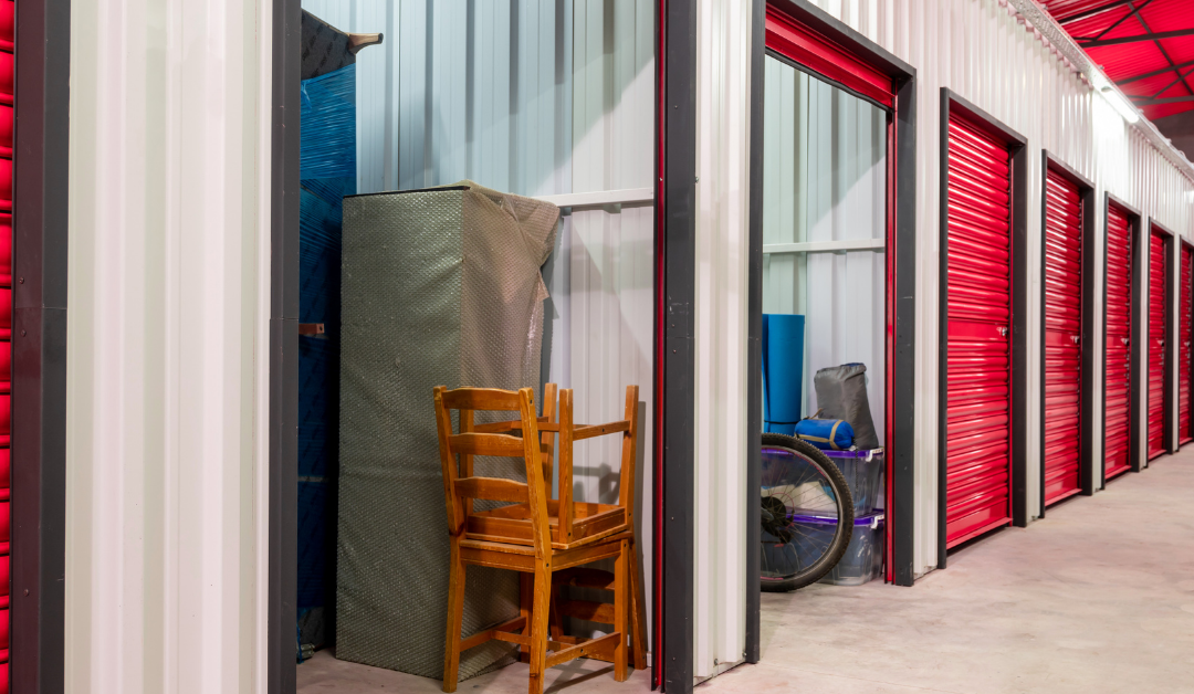 A row of red self-storage units with one unit open, revealing a mattress, chair, and other belongings inside.