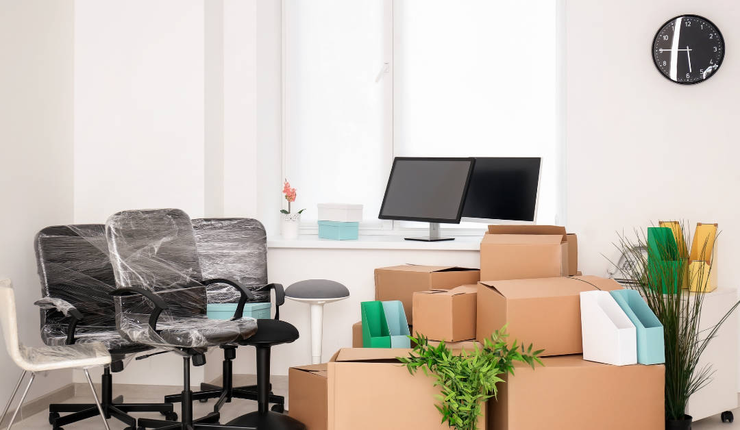 An office space in the middle of a move. Chairs are wrapped in plastic, boxes are stacked, and office equipment is packed.