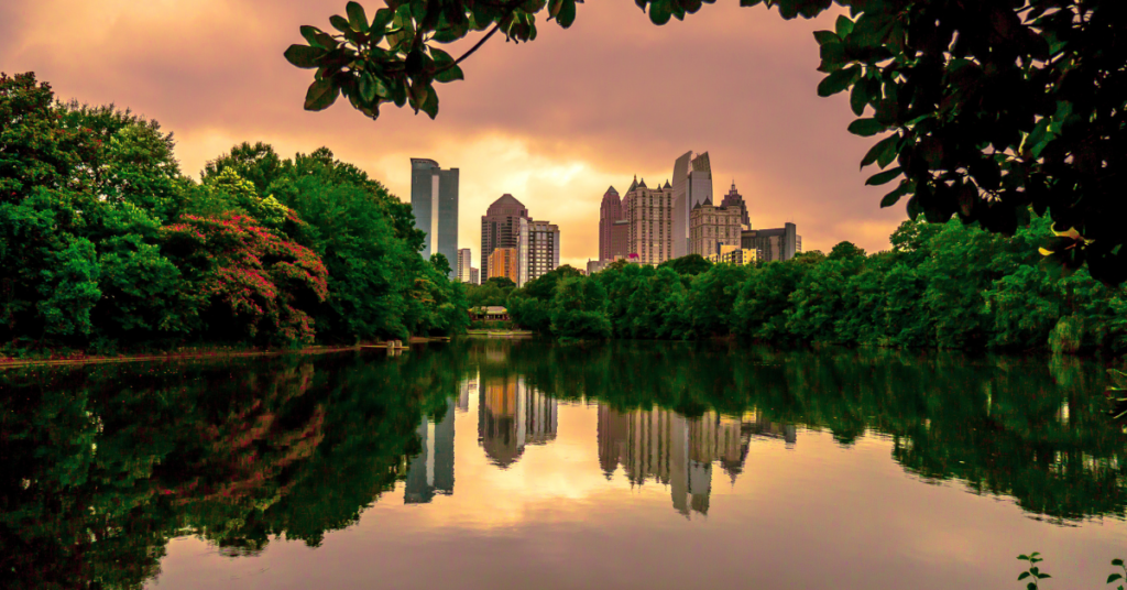 A scenic view of a lake surrounded by trees in a park, with a cityscape reflected in the water. The sky is a vibrant orange and pink, suggesting it's sunrise or sunset.